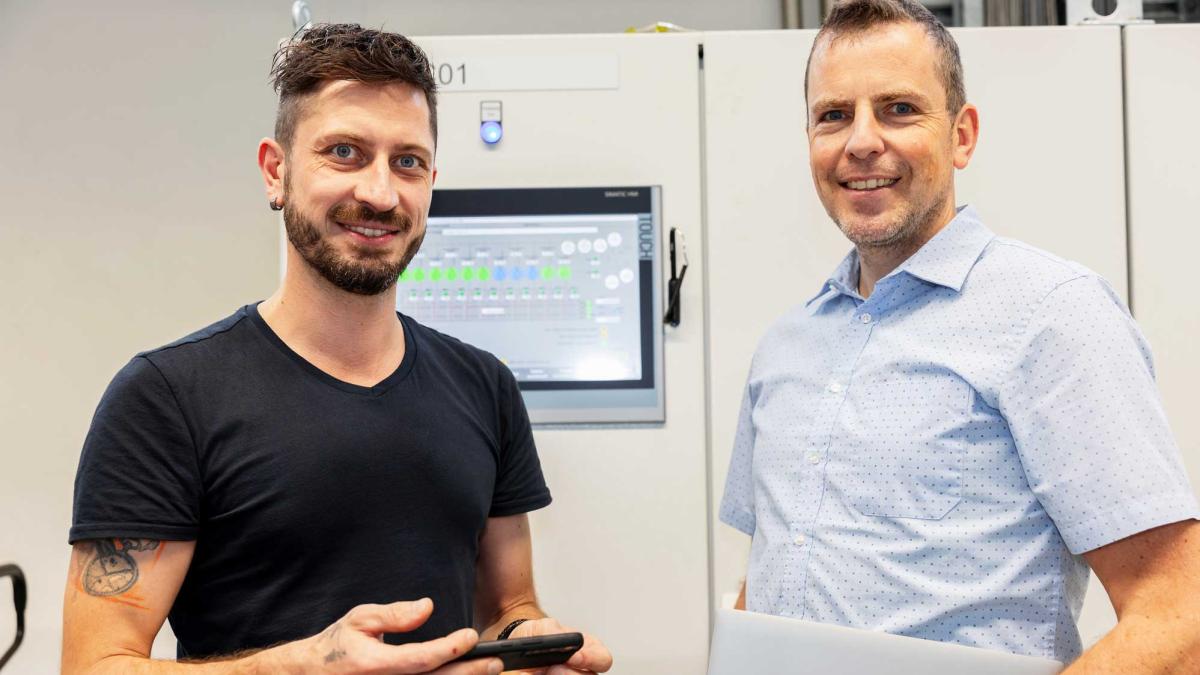 Two men standing next to each other in front of a machine control panel and looking into the camera