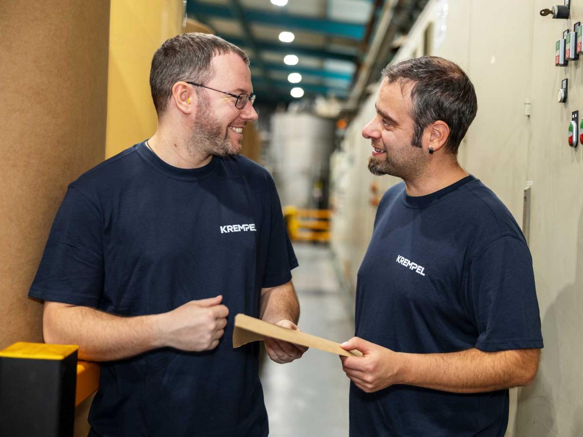 Two men in blue shirts standing in a production hall lookint at each other and talking